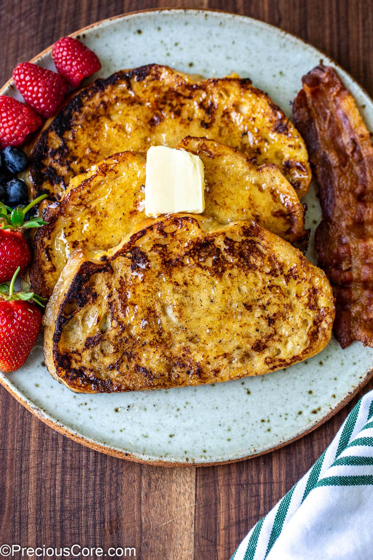 3 slices of sourdough French toast on a plate with fruit and bacon on the side.