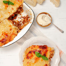 Slice of Lasagna on a plate with a bowl of freshly baked lasagna on the side.